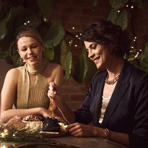Manuka Health Manuka Honey Kit. Two women sitting at a table with a decorative setting, smiling and interacting.