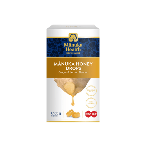 A box of Mânuka Health Mânuka Honey Drops (Ginger & Lemon Flavour) on a solid white background. The packaging is white with a dark blue band at the top, a gold banner, and an illustration of honey dripping from a piece of ginger root.