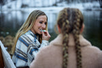 Two women sitting by a lake, one applying makeup to the other's face.